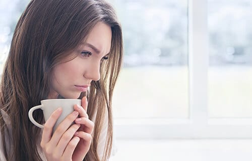 A woman holding a mug of coffee, looking distressed. She's thinking about attending Depression and Addiction treatment at Crest View.
