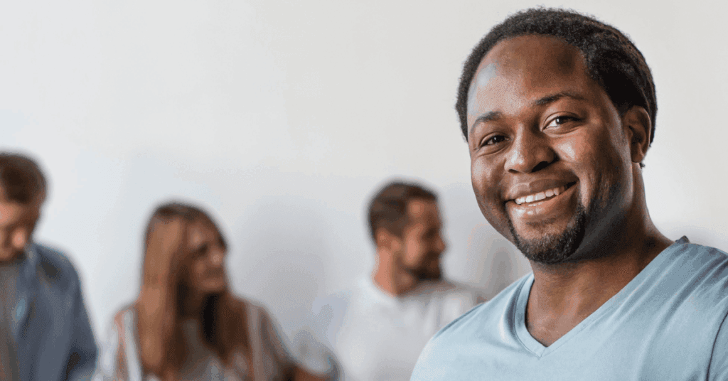 A sober man in a light blue shirt smiles at the camera, while three people in the background talk and interact with each other in a blurred setting.