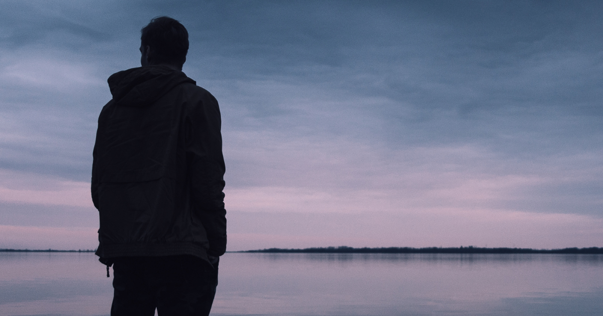 A silhouette of a somber man stands in front of a lake, contemplating. Trauma, addiction, and mental health are all intertwined.