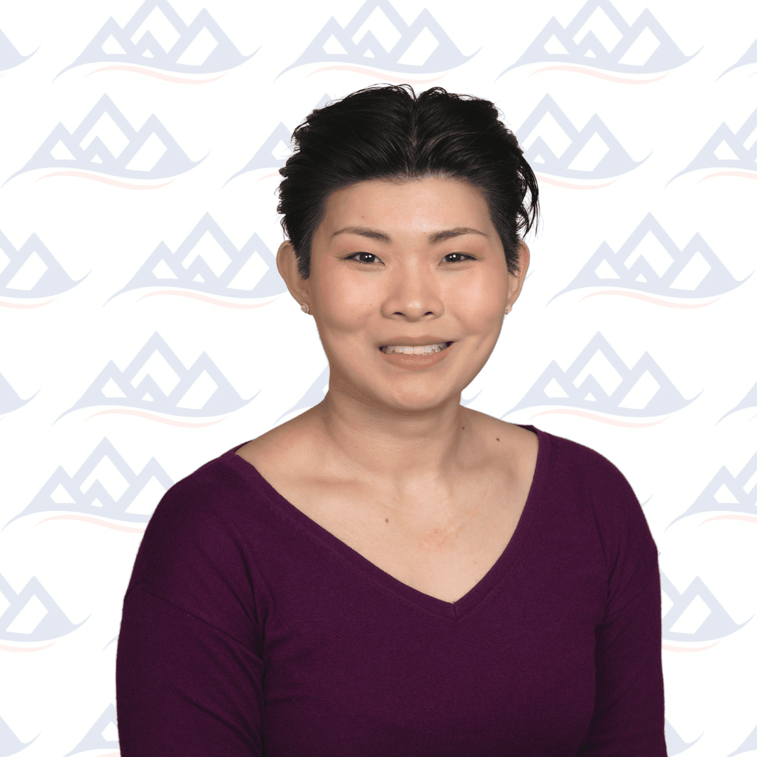 A certified addiction professional with short, dark hair smiles at the camera. They wear a deep purple V-neck top and sit in front of a white background featuring a repeating pattern of light blue abstract mountain shapes.