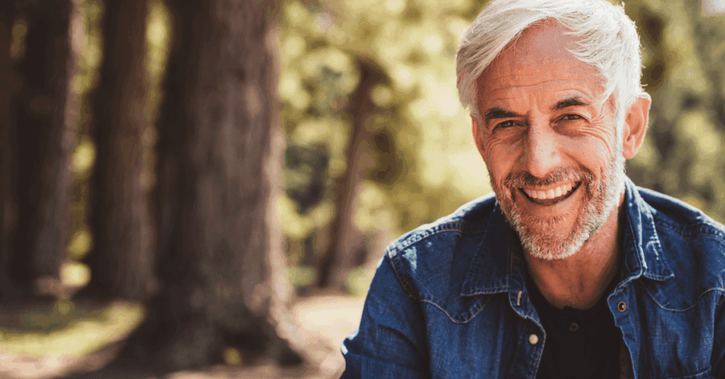 An older, white-haired man, in recovery from alcohol use disorder, looks to camera with an open mouth smile, with a woodsy scene in the background.