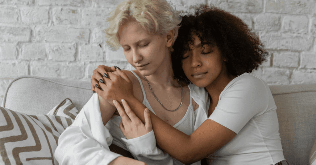 A female couple from Crest View's LGBTQIA+ program sit in a supportive embrace with their eyes closed on a tan couch with a white brick background.