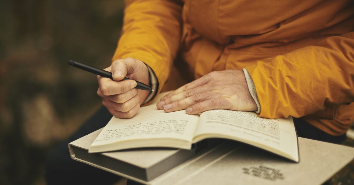 Sober Gratitude Practices A person wearing an orange jacket writes in a notebook with a pen while resting it on their lap. Another hand holds the notebook open, and some handwritten notes are visible on the page, showing the man working on sober gratitude practices.