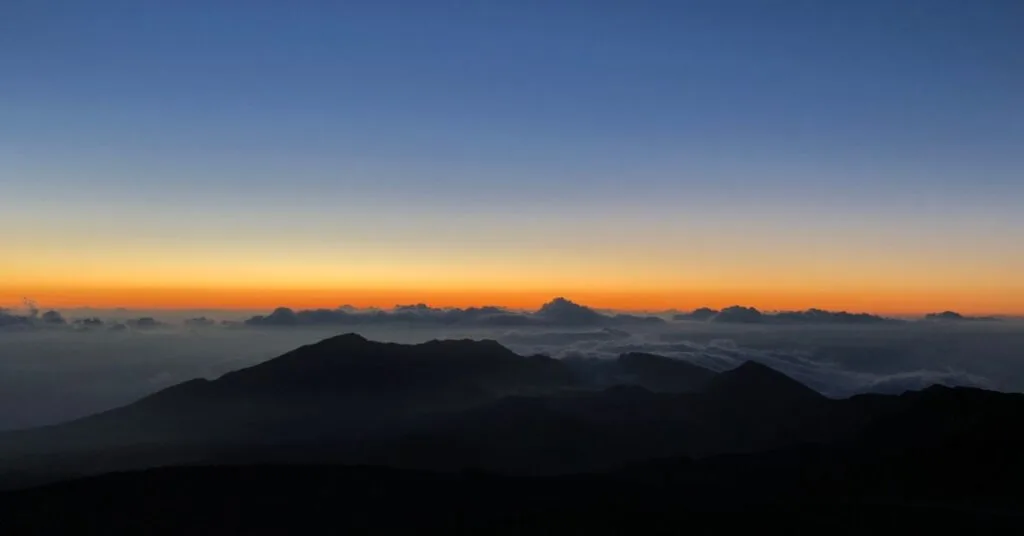 A silhouette of mountain peaks under a clear sky at sunrise or sunset, with a band of orange and yellow light on the horizon and clouds resting below the mountains, representing Holidays in addiction recovery.