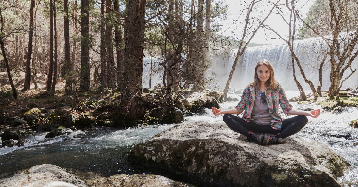 A woman sits cross-legged on a large rock by a flowing river, meditating in a forest with a waterfall in the background, showing one of many relapse prevention strategies. Sunlight filters through the trees.