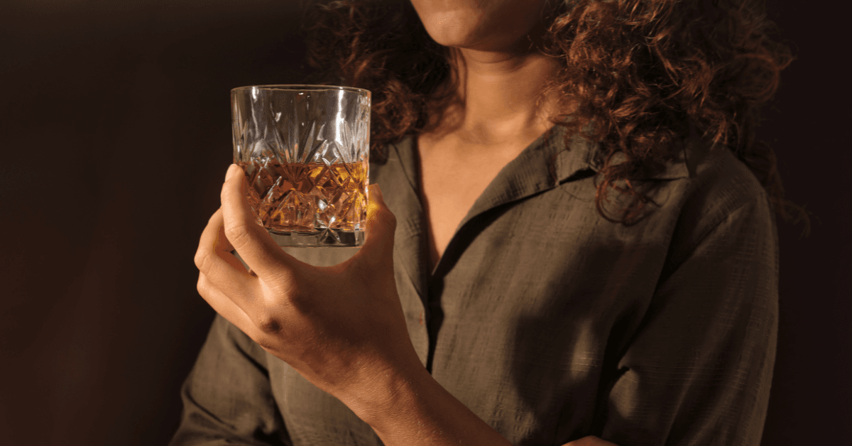 Individual with curly hair holds a glass of whiskey, brown shirt visible—signs of alcohol use disorder shown.