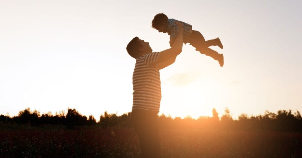 A father in a striped shirt lifts his child at sunset in a field, symbolizing hope, healing, and dads in recovery from addiction.