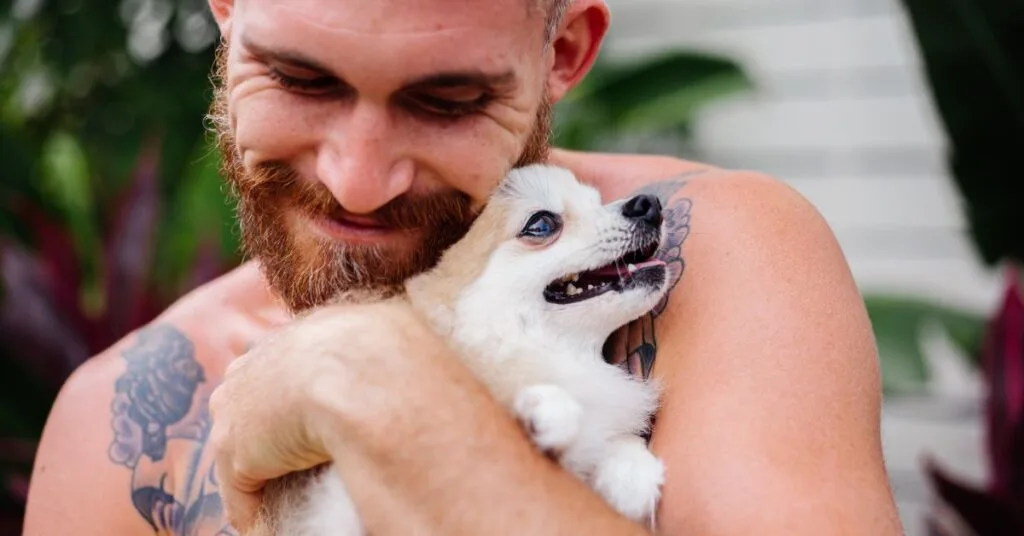 A bearded man with tattoos hugs a happy puppy outdoors, symbolizing recovery, support, and healing at Crest View Recovery Center, one of the rehabs that allows dogs.