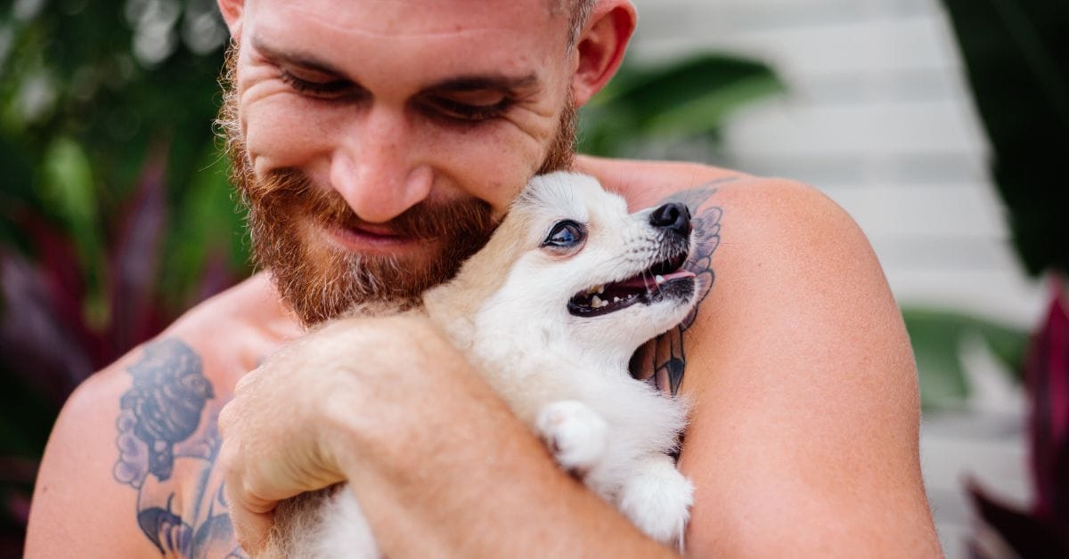 A bearded man with tattoos hugs a happy puppy outdoors, symbolizing recovery, support, and healing at Crest View Recovery Center, one of the rehabs that allows dogs.