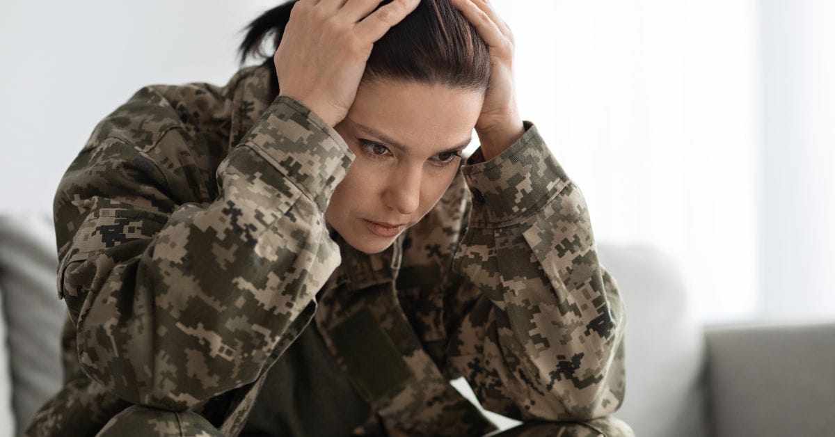 A person in military camouflage sits with head in hands, showing stress—veteran struggling with addiction, needing a veteran-focused addiction rehab.