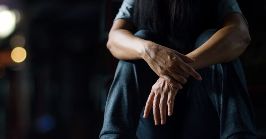 Person sitting with arms crossed over knees and head down, reflecting addiction recovery struggles in a dimly lit treatment setting for PTSD awareness month.