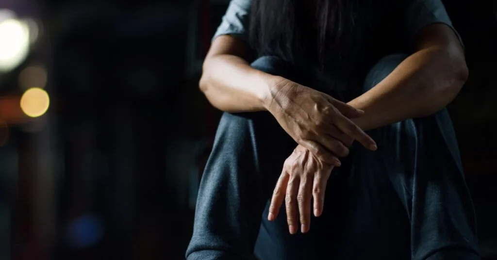 Person sitting with arms crossed over knees and head down, reflecting addiction recovery struggles in a dimly lit treatment setting for PTSD awareness month.