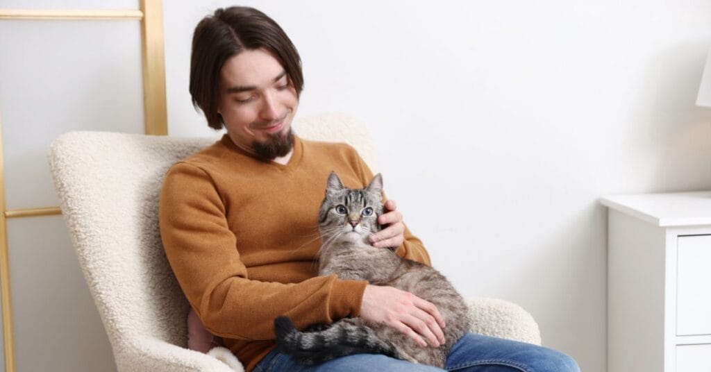 A man sits comfortably in a cozy chair at home, smiling and holding a grey tabby cat on his lap. The serene, supportive setting reflects the benefits of pets for addiction recovery.