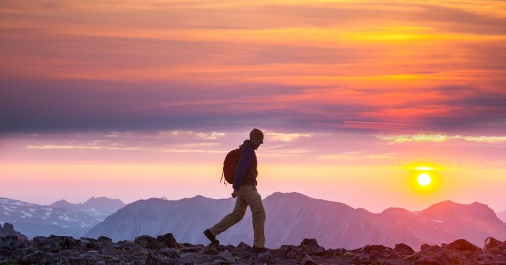 A person with a backpack hikes along a rocky mountain ridge at sunset, showing one of the addiction recovery activities Crest View Recovery uses.